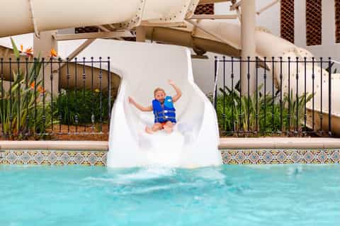 Child sliding down white water slide into turquoise pool at resort