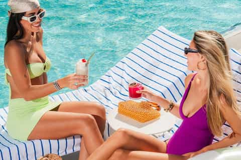 Two women in swimwear enjoying cocktails by a crystal-clear turquoise pool with snacks