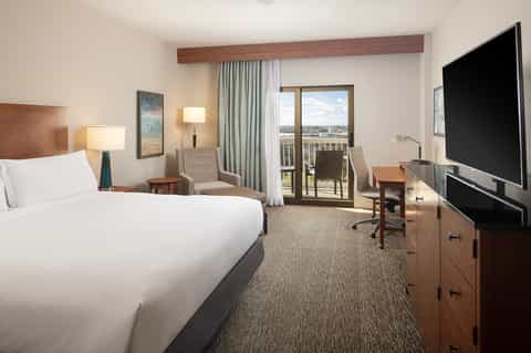 Hotel bedroom with queen bed, balcony overlooking waterfront, sitting area, TV, and modern furnishings