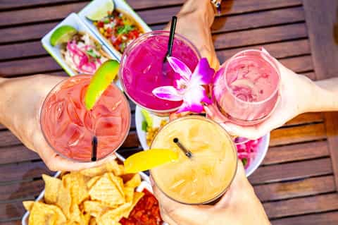 Colorful tropical cocktails held by person poolside with appetizers and flowers