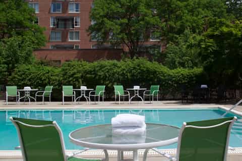 Urban resort pool with green lounge chairs, brick building, and lush tree backdrop