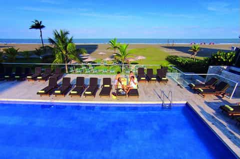 Resort pool overlooking beach with lounge chairs, palms, and ocean view during daytime