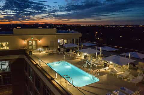 Rooftop pool at night with illuminated water, lounge chairs, and city lights