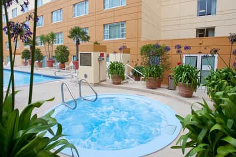 Hot tub surrounded by potted plants with resort building and larger pool visible in background courtyard