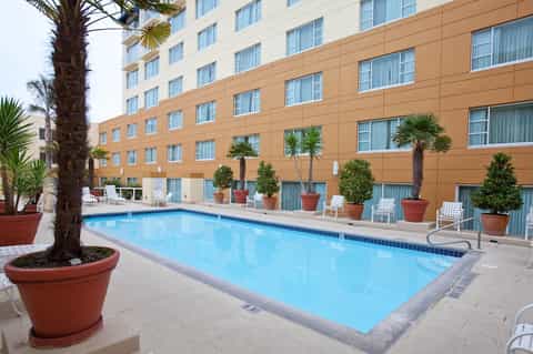 Outdoor swimming pool with tan hotel building facade, palm trees, and white lounge chairs