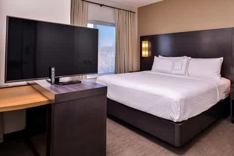 Modern hotel bedroom with king bed, dark headboard, large TV, and city view from window