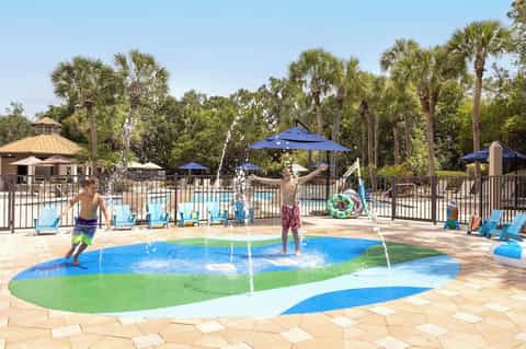 Family splash pad and pool area with colorful interactive water features surrounded by palm trees