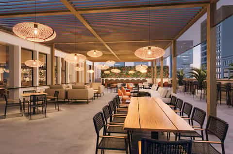 Modern rooftop terrace lounge with woven pendant lights, wooden long table, and urban cityscape in background