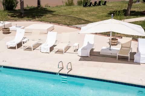 Resort pool deck with white lounge chairs, umbrellas, fire pit, and manicured lawn with trees
