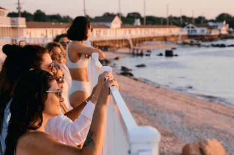 People enjoying sunset from a beach boardwalk railing overlooking water and harbor
