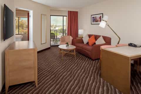 Hotel suite with balcony view, brown sofa, work desk, and television entertainment