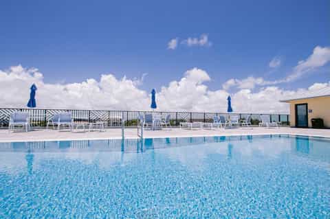 Resort pool with white loungers, blue umbrellas, and ocean views beyond railing