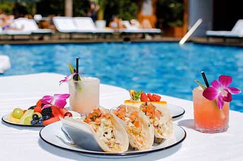 Poolside dining spread with tacos, tropical fruit plate, and cocktails garnished with pink orchid flowers