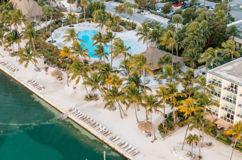 Aerial view of beachfront resort with turquoise pool, palm trees, sandy beach, and waterfront docks