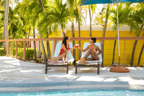 Two women relaxing on benches by a pool with yellow building, palm trees, and turquoise shade umbrella
