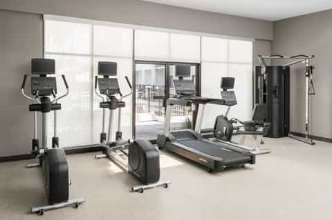 Bright fitness center with cardio equipment, treadmills, ellipticals, and large windows overlooking a balcony