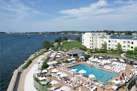 Waterfront resort pool with white umbrellas, loungers, and multi-story white building overlooking bay