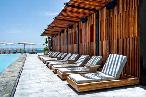 Resort poolside cabana lounge with striped cushioned beds and wooden shade pergola by water