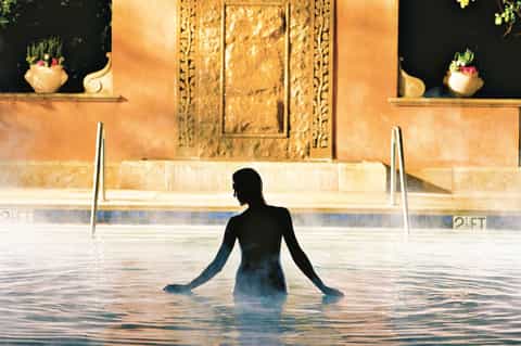 Woman in dark swimsuit relaxing in a steaming thermal pool with ornate golden temple architecture backdrop