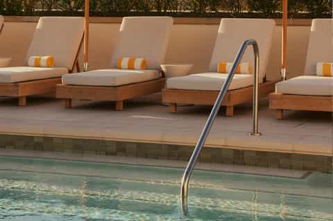 Pool deck with wooden lounge chairs, cream cushions, and metal railing beside turquoise water