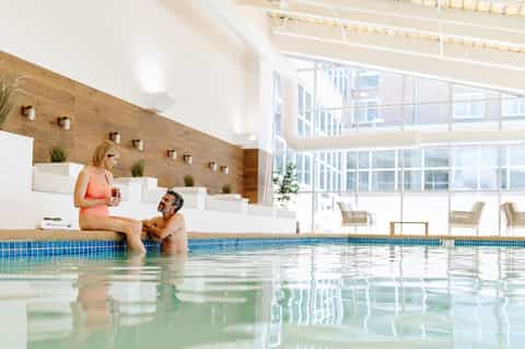 Indoor pool area with couple at pool edge, wood-paneled walls, skylights, and lounge seating