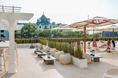 Waterfront lounge area with wooden pergolas, woven seating, grasses, and coastal mansion in background