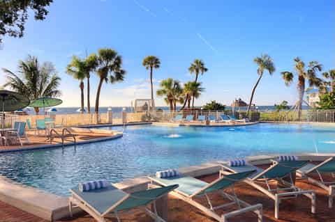 Resort outdoor pool area with lounge chairs, palm trees, ocean view, wooden deck, and clear blue water
