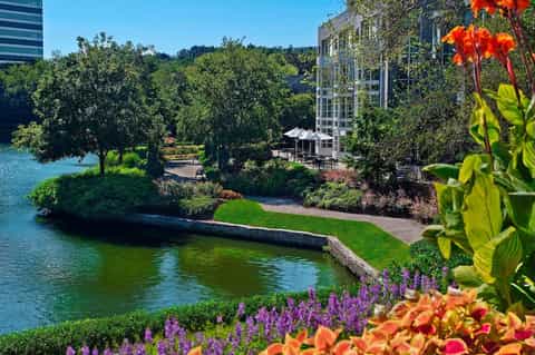 Waterfront hotel building overlooking river with lush trees, colorful flower gardens, and green manicured landscape