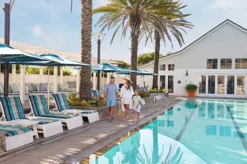Family walking along outdoor pool deck with loungers, palm trees, and modern building