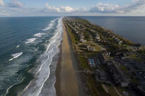Aerial view of coastal peninsula with sandy beach, ocean waves, homes, and waterfront resort facilities
