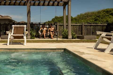 Two guests on porch swing overlooking small pool with wooden pergola and beach landscaping
