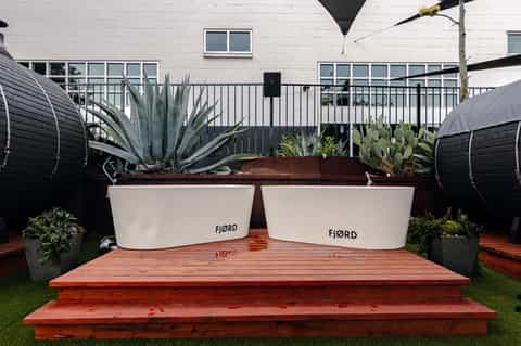 Outdoor hot tub lounge area with white FJORD branding, agave plants, red deck, and modern hotel building.