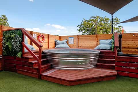 Rustic hot tub with red wooden deck, cushioned seating, and shade canopy in natural outdoor setting