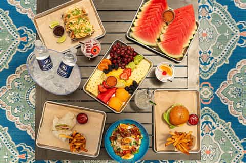 Overhead view of resort poolside dining spread with fresh fruit, tacos, burgers, and beverages on colorful rug