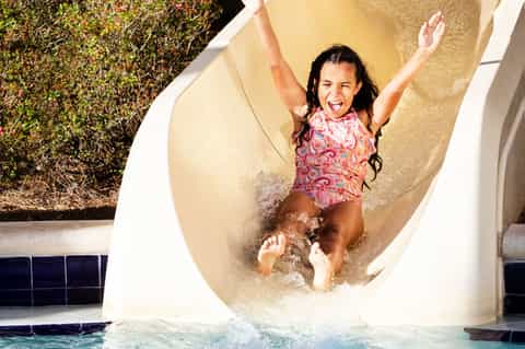 Child sliding down water slide into pool wearing pink swimsuit with arms raised
