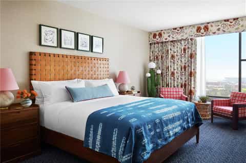 Guest bedroom with orange woven headboard, white bedding, blue throw blanket, floral curtains, and city view