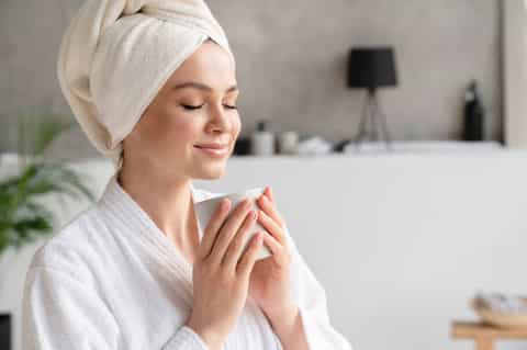 Woman in white spa robe and towel wrap holding warm beverage in modern bathroom setting