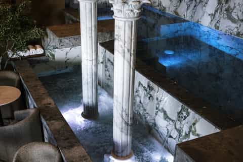 Indoor thermal hot spring bath with blue water, white columns, and stone seating areas