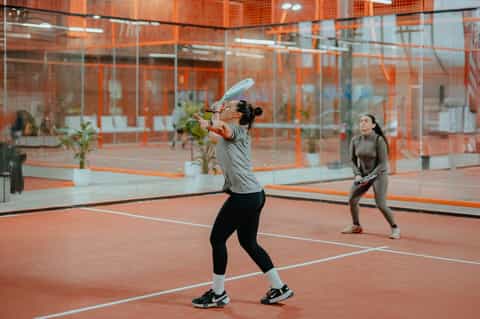 Indoor tennis court with red clay and orange glass enclosure, two players