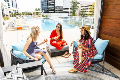 Three women enjoying cocktails on a rooftop lounge with a resort pool and city skyline in background
