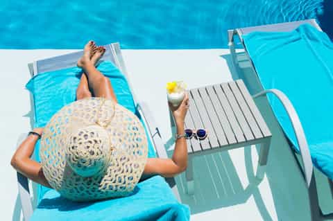 Overhead view of resort guest lounging by bright blue pool with hat and tropical cocktail