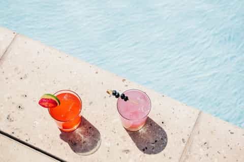 Two colorful cocktails on pool deck edge with clear water and lime garnish