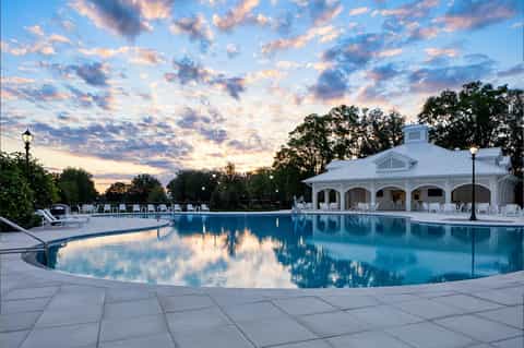 Elegant resort pool at dusk with illuminated pool house, white lounge chairs, and sunset sky reflections