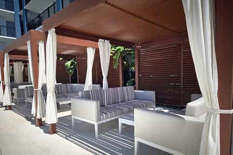 Covered outdoor lounge area with wooden pergola, white curtains, striped seating, and tropical plants