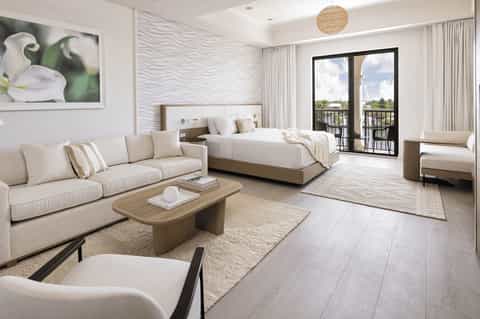 Spacious suite with cream sofa, king bed, wave-textured walls, and balcony overlooking grounds