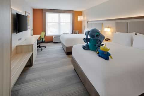 Colorful family hotel room with two queen beds, orange accent wall, TV, desk, and Disney character plush toys
