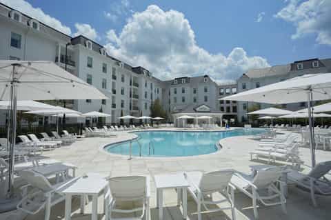 Curved resort pool surrounded by white loungers, umbrellas, stone patio, and colonial-style hotel building