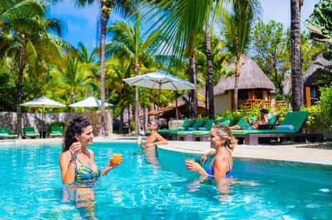 Resort pool scene with guests swimming, palm trees, white umbrellas, and thatched-roof cabanas