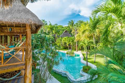 Tropical resort grounds featuring thatched-roof bungalows, turquoise pool, and lush vegetation