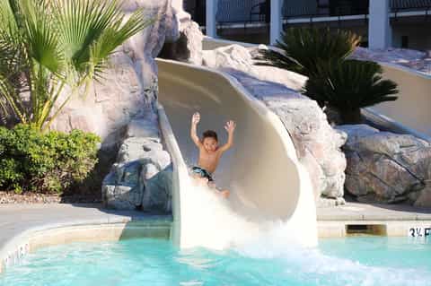 Child sliding down white water slide into turquoise pool surrounded by white rock features and palm trees
